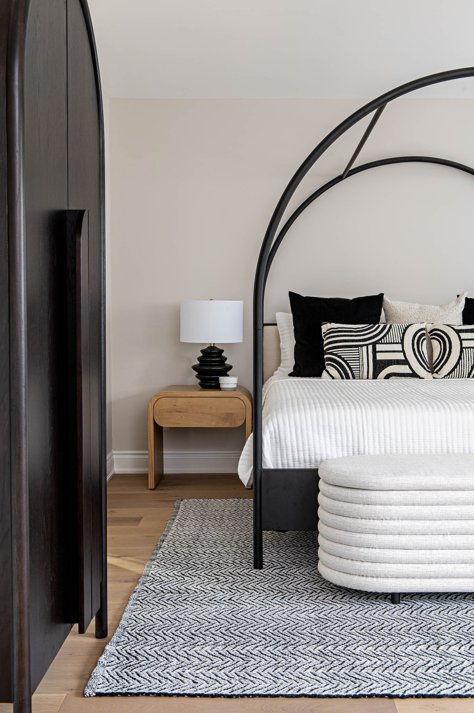 Image of modern Primary Bedroom, black metal arched canopy bed with white bedding and black and white pillows, natural wood side tables, black table lamps, black and white area rug, upholstered bench at foot of bed.