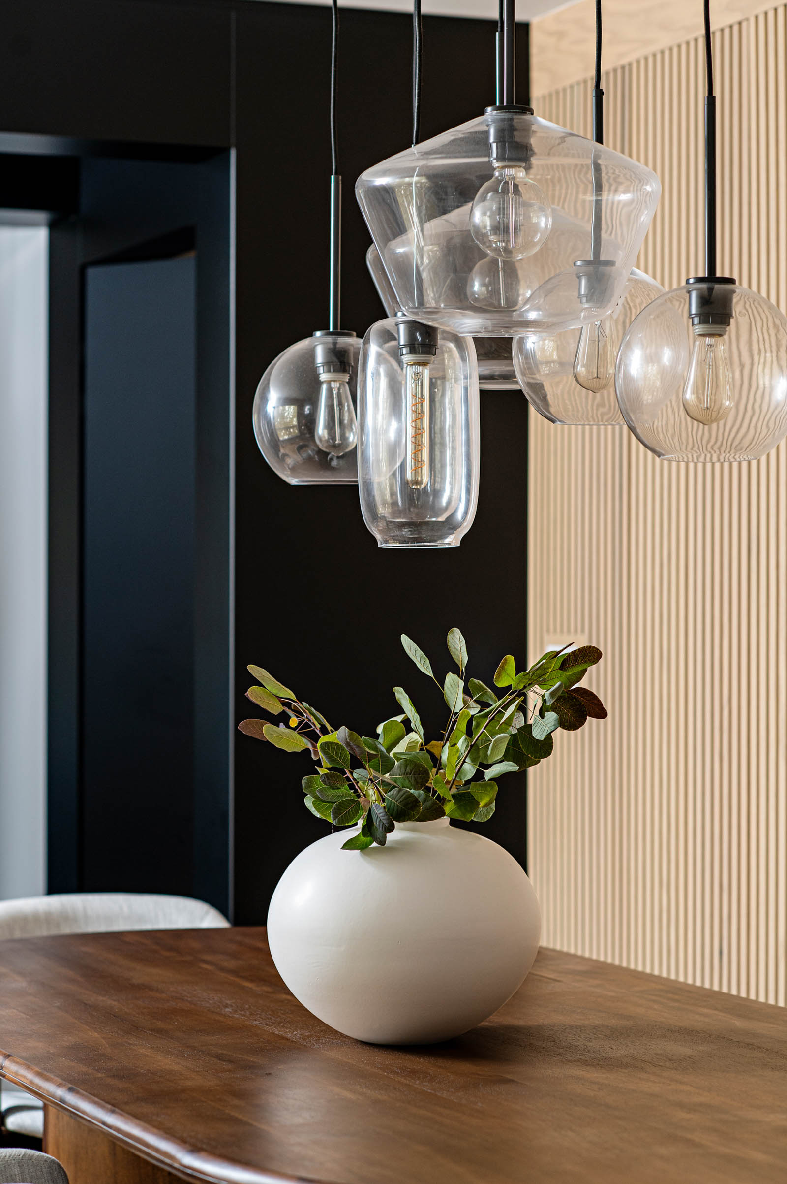 Image of Dining Room detail of clustered glass pendant lighting against a natural oak reeded wall. White centerpiece bowl with green foliage on table.