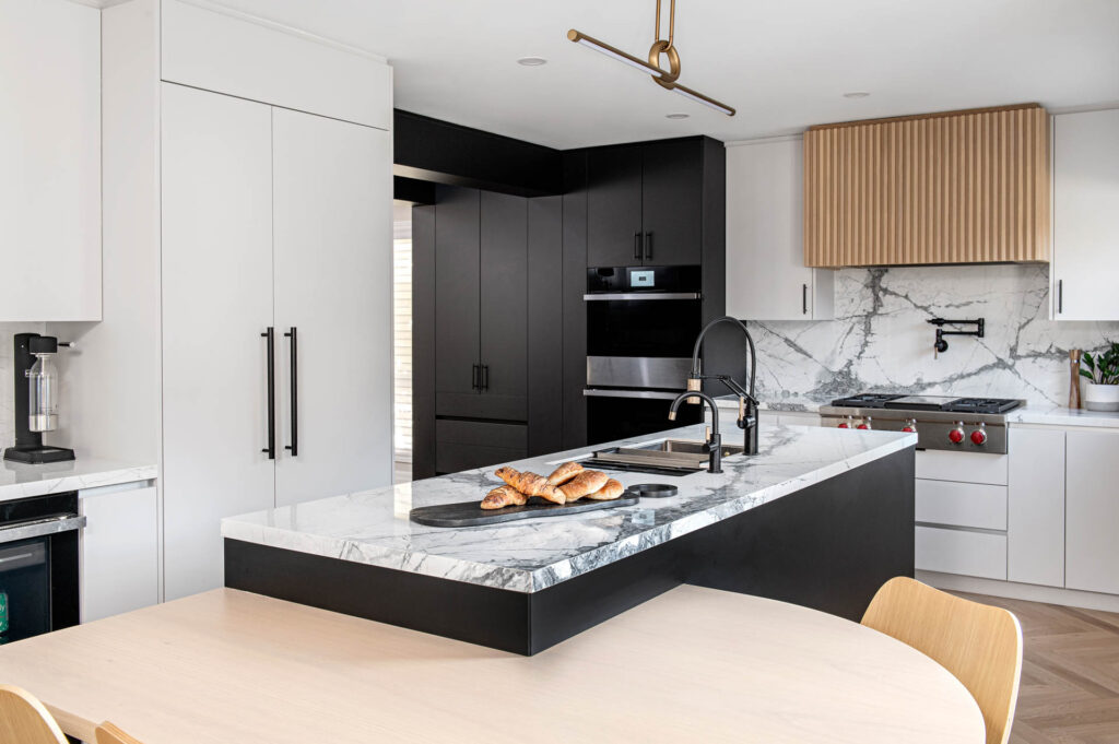 Image of a completed Modern kitchen renovation with marble look porcelain countertop and Backsplash, light grey slab cabinets and drawers, Natural Wood Reeded hood fan, wall ovens, integrated paneled fridge, Large island with cantilevered table.