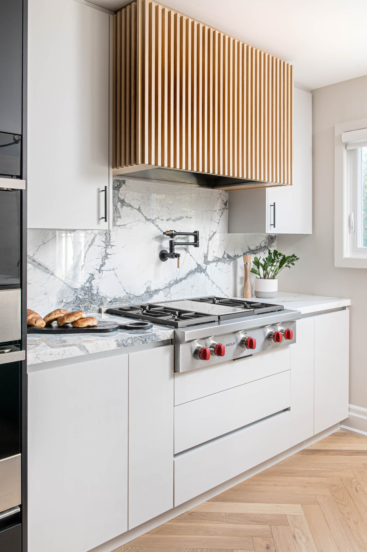 Image of Contemporary kitchen with marble look porcelain countertop and Backsplash, light grey slab cabinets and drawers, Natural Wood Reeded hood fan, Wolf Rangetop, Black Wall Mount Pot Filler.