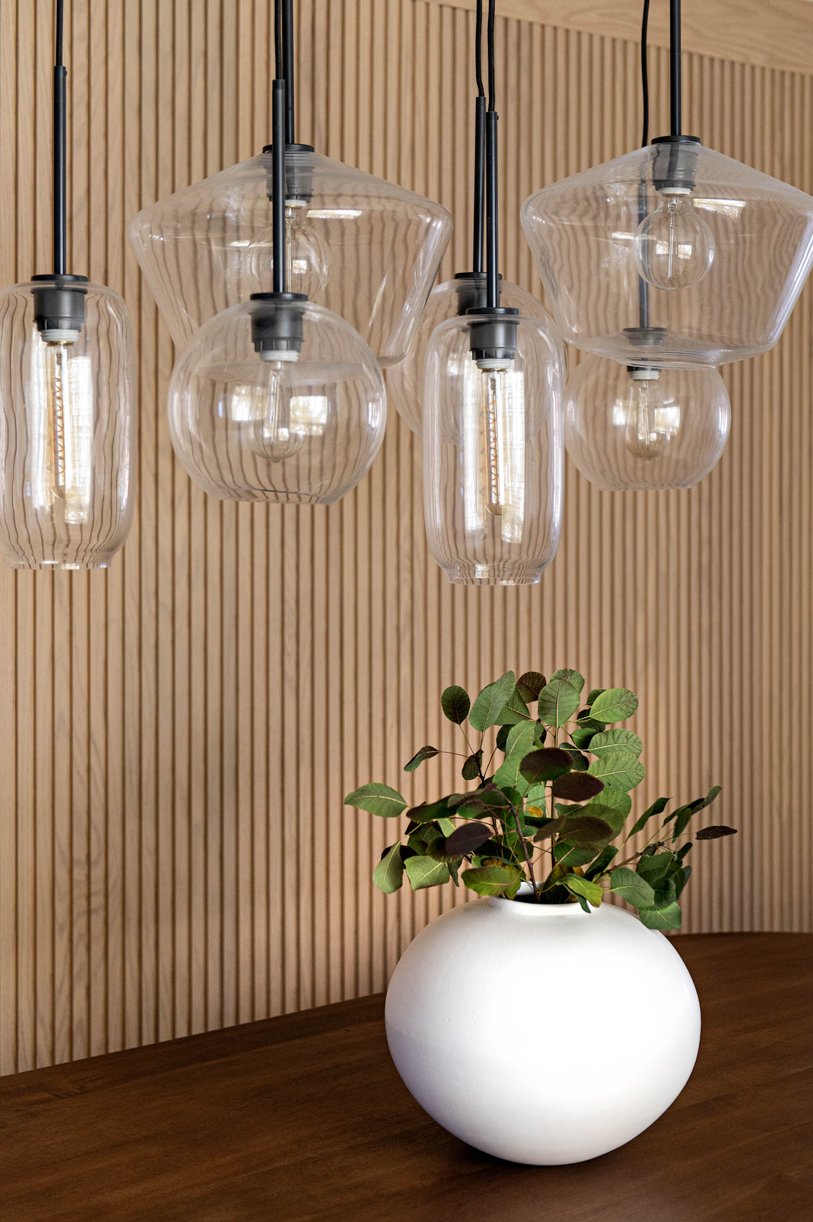 Image of Dining Room detail of clustered glass pendant lighting against a natural oak reeded wall. White centerpiece bowl with green foliage on table.