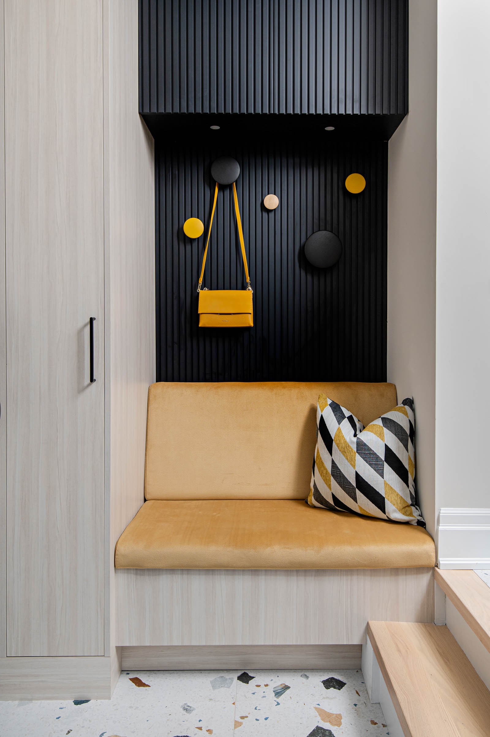 Image of modern mudroom, showcasing custom storage, black reeded wall detail with round Muuto wall hooks, custom bench with gold fabric, and colour large scale terrazzo floor.