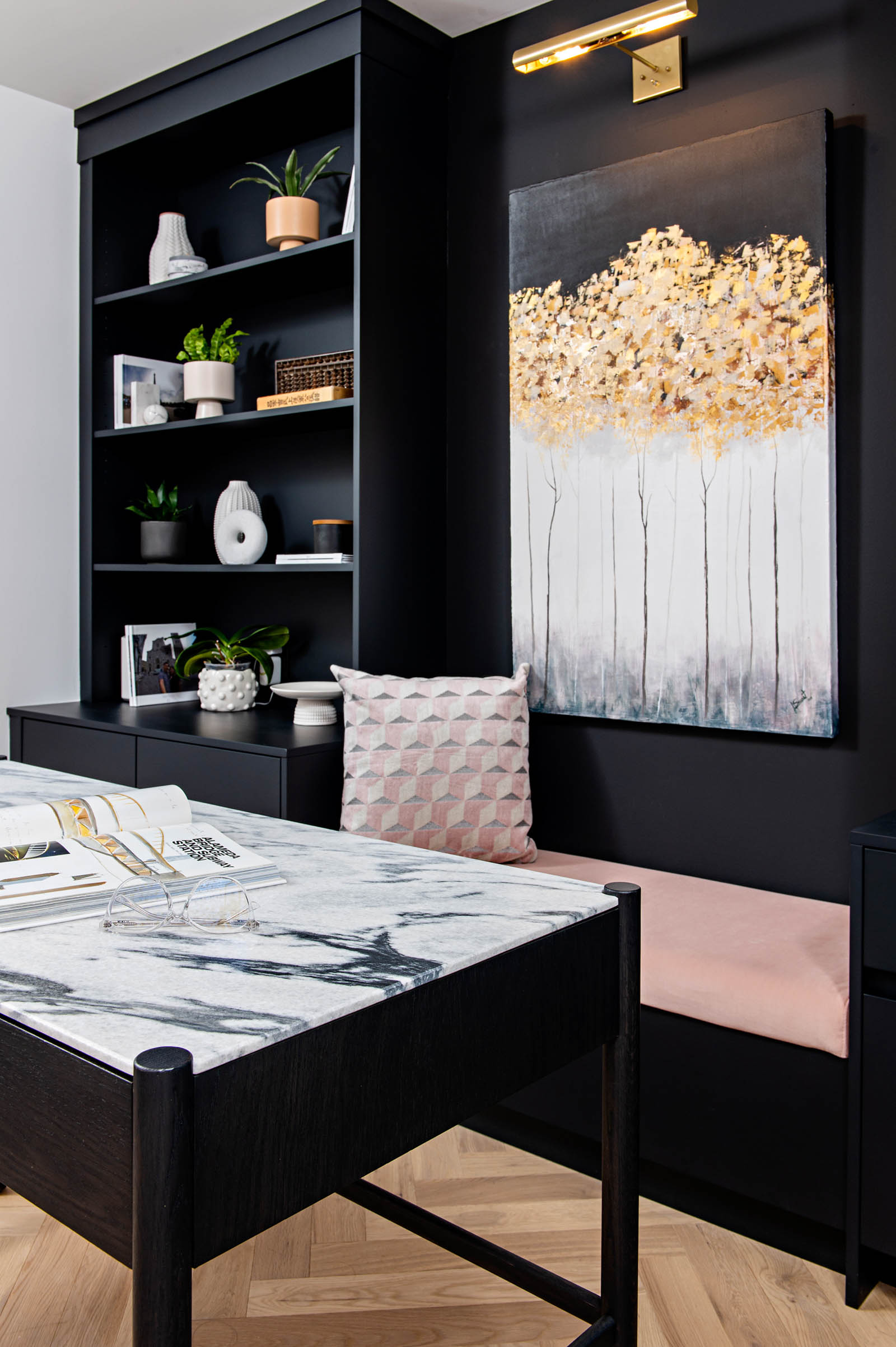 Image of custom built home office black shelving, beside it a bench with upholstered pink fabric and artwork above. modern desk with stone top.