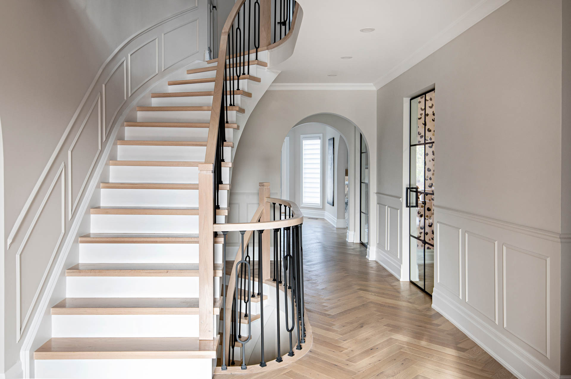 Image of home entrance with renovated staircase that has black iron pickets, natural oak railing, white risers and oak steps. flooring is a light oak engineered hardwood herringbone.