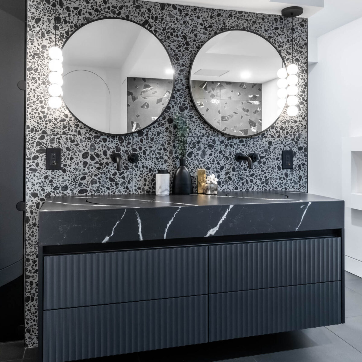 Image of a contemporary primary ensuite bathroom, showcasing a sleek black floating vanity with fluted detail and black with white vein quartz countertop, integrated sink, terrazzo porcelain wall tile, round vanity mirror and wall mounted black faucet.