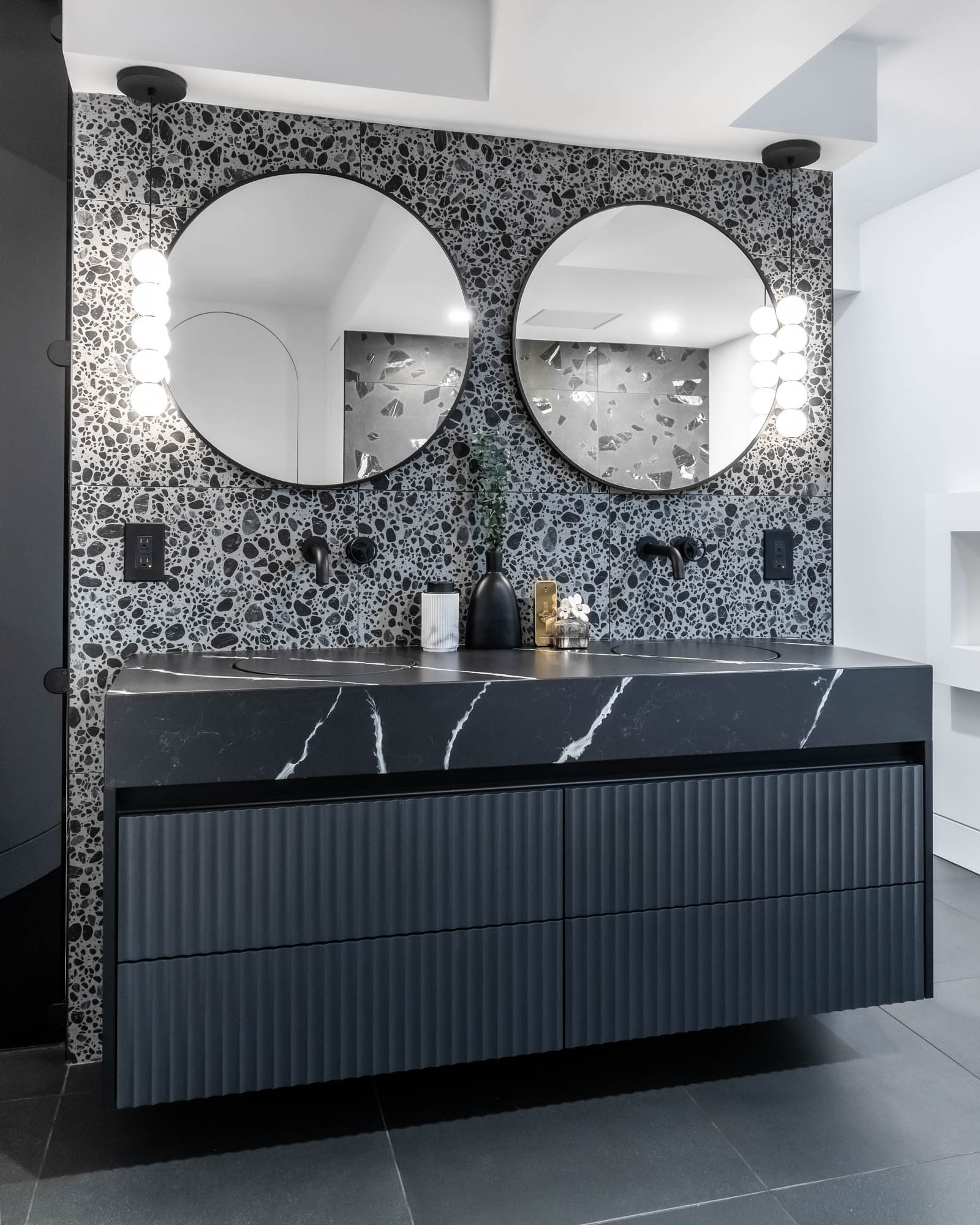 Image of a contemporary primary ensuite bathroom, showcasing a sleek black floating vanity with fluted detail and black with white vein quartz countertop, integrated sink, terrazzo porcelain wall tile, round vanity mirror and wall mounted black faucet.