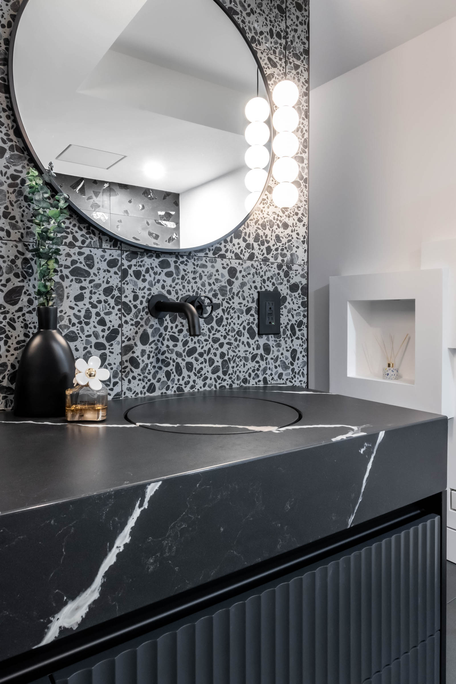 Closeup image of a contemporary primary ensuite bathroom, showcasing a sleek black floating vanity with fluted detail and black quartz countertop, contemporary integrated sink, terrazzo porcelain tile, and wall mounted black faucet.