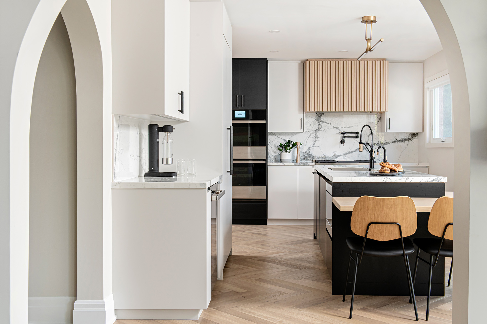 Image of Modern kitchen renovation with marble look porcelain countertop and Backsplash, light grey slab cabinets and drawers, Natural Wood Reeded hood fan, wall ovens, integrated paneled fridge, Large island with cantilevered table.