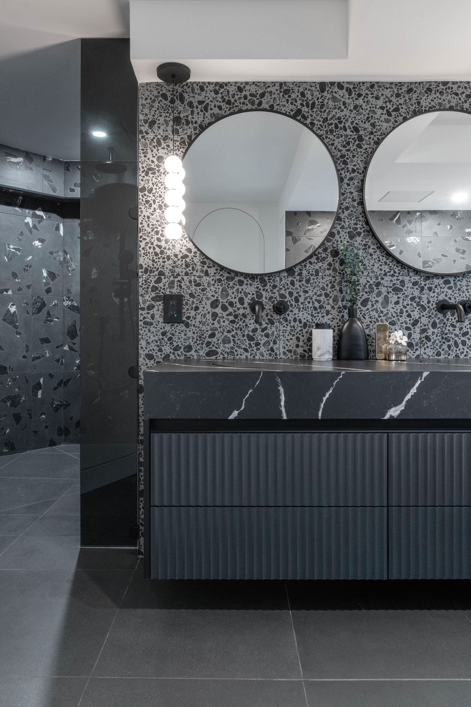 A modern bathroom vanity with a black quartz countertop, terrazzo backsplash, and round mirrors, with a glimpse of the shower area on the left.