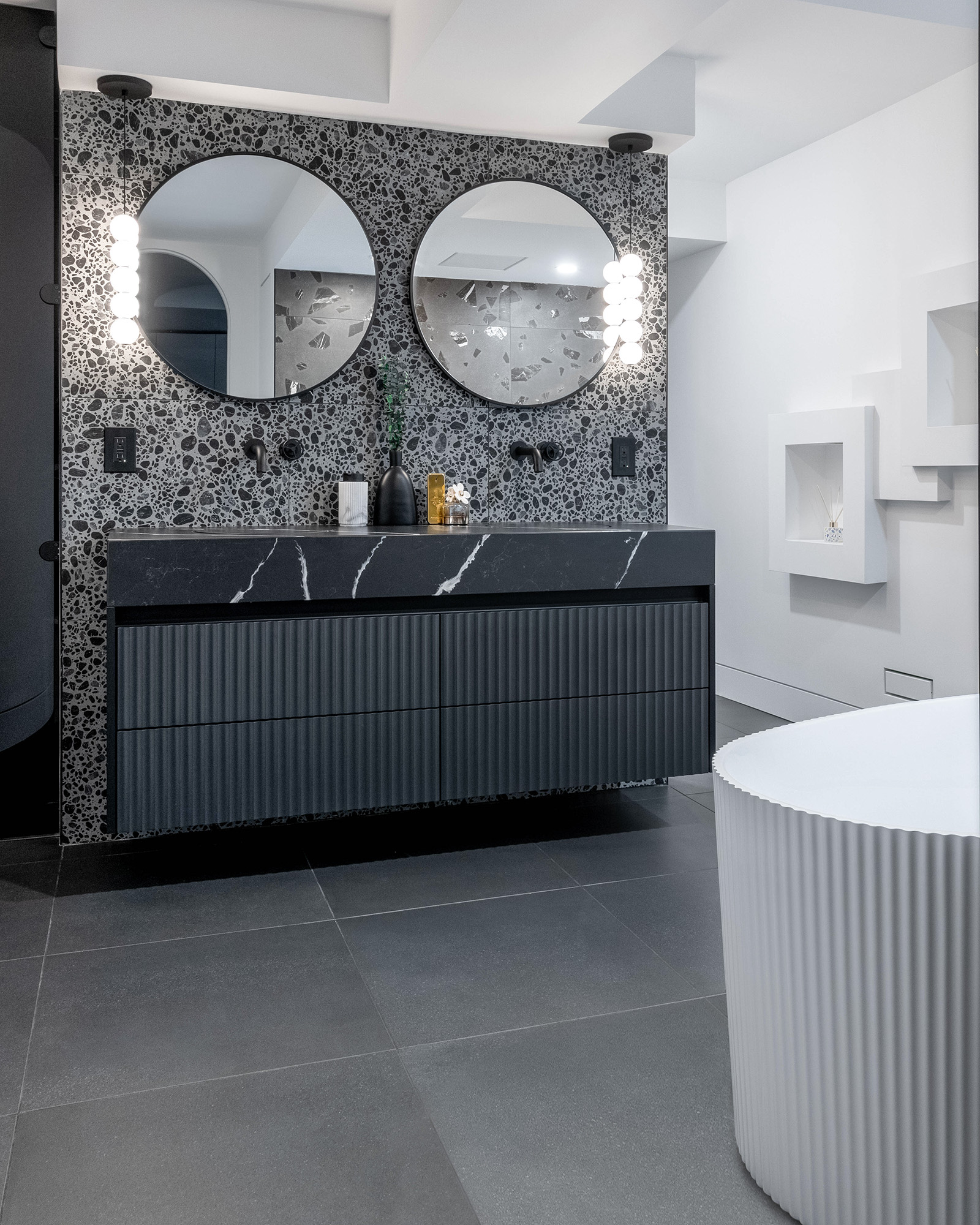 Modern bathroom with a double vanity, large-scale terrazzo porcelain tile wall, grey porcelain tile floor, and black wall-mounted faucets.