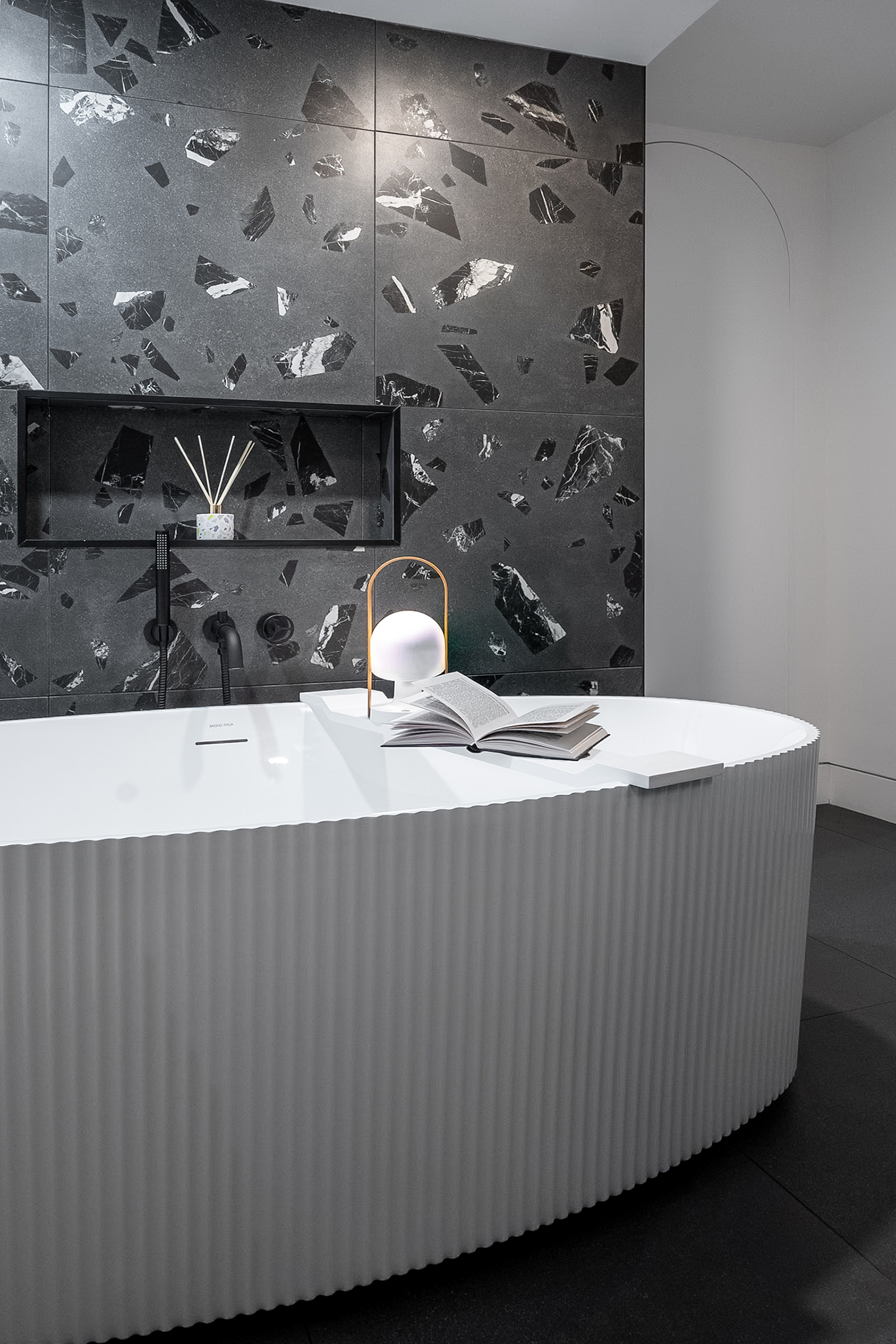 Large-scale terrazzo porcelain tile wall with a grey porcelain tile floor and a white ribbed freestanding tub with a niche