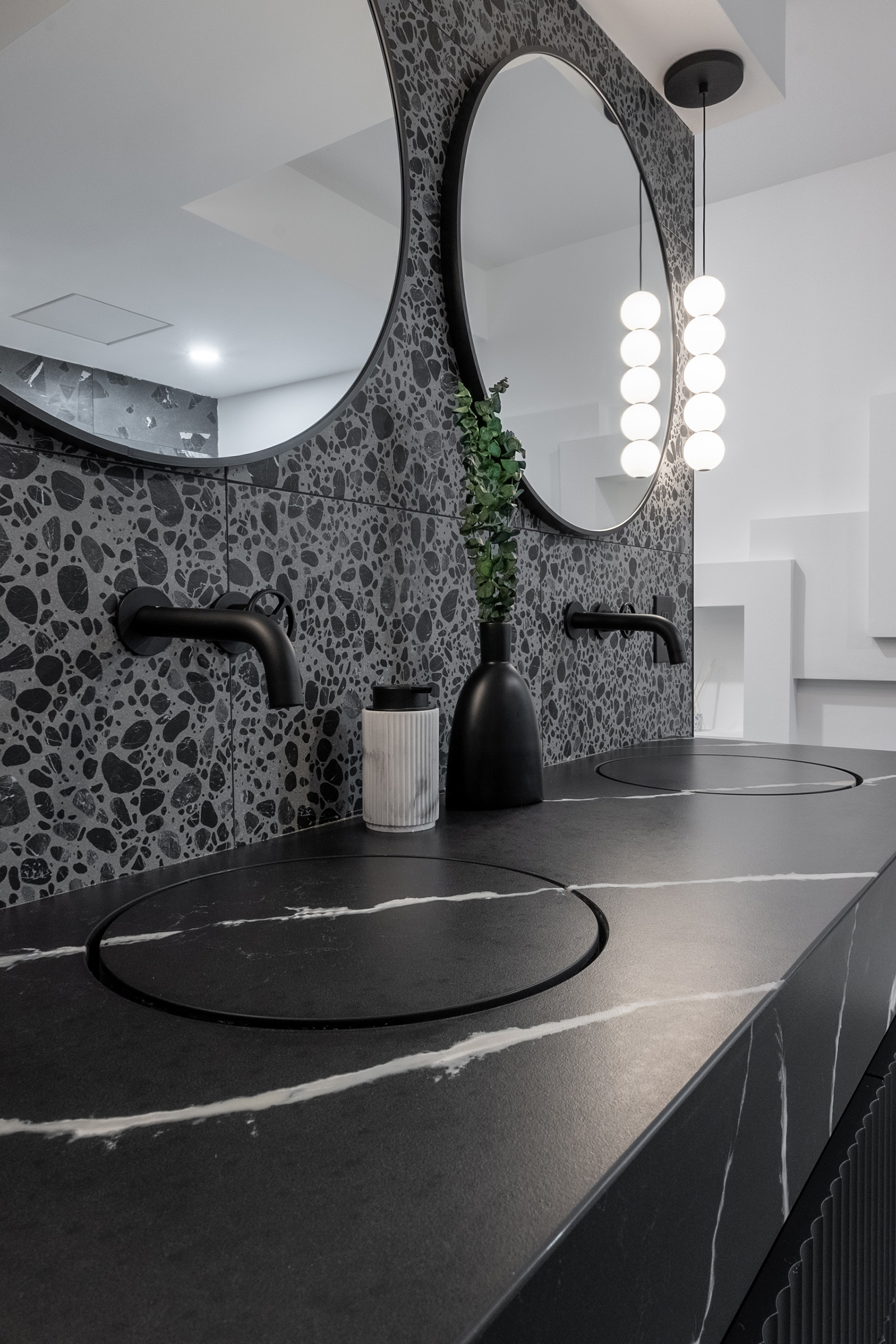 A close-up view of a modern bathroom vanity featuring a flat washbasin sink with a black quartz countertop and terrazzo backsplash, accented by round mirrors and minimalist black fixtures.