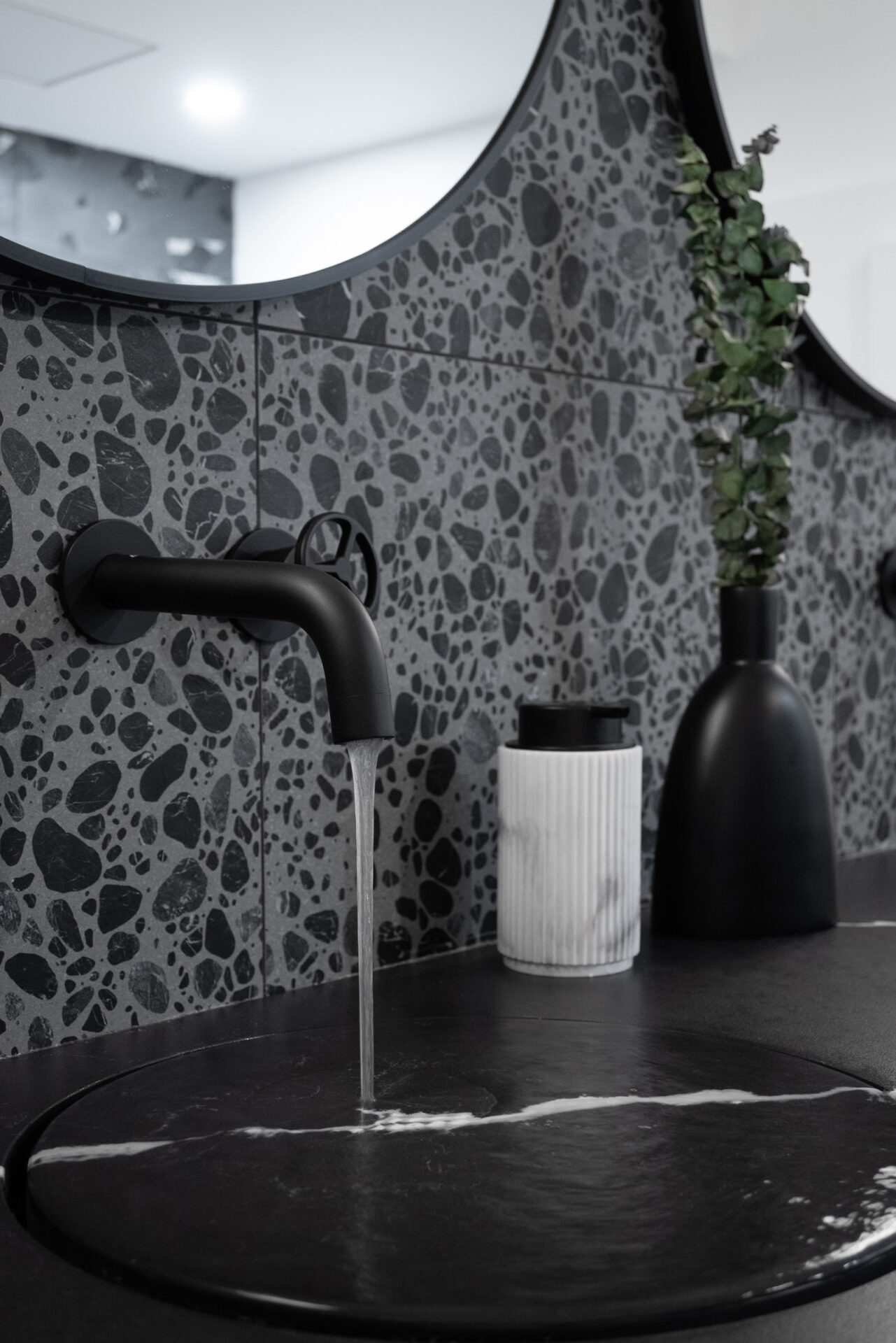 A close-up view of a modern bathroom sink with water flowing from a matte black faucet into a black quartz countertop integrated sink, set against a terrazzo tile backsplash.